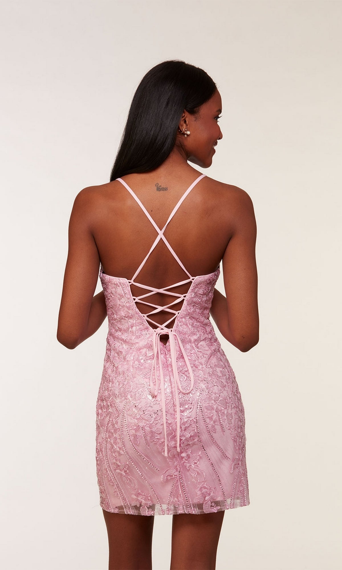 A woman stands facing away from the camera against a plain background, wearing the Beaded Embroidered Short Homecoming Dress 4615 in light pink with thin straps and a lace-up back.